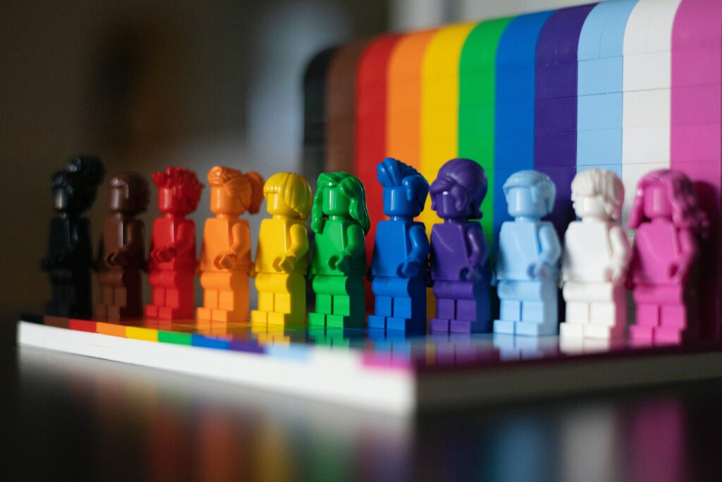 Legofiguren, jeweils einfarbig, in den Farben der Progress-Pride-Flag, im Hintergrund wiederholen sich die Farben als Streifen